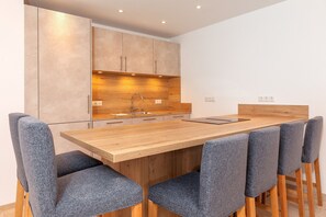 A modern kitchen with wooden cabinets, a central island with a sink, and a dining area with a wooden table and grey upholstered chairs. - Vacation apartment living on the Lofer (Reit im Winkl)