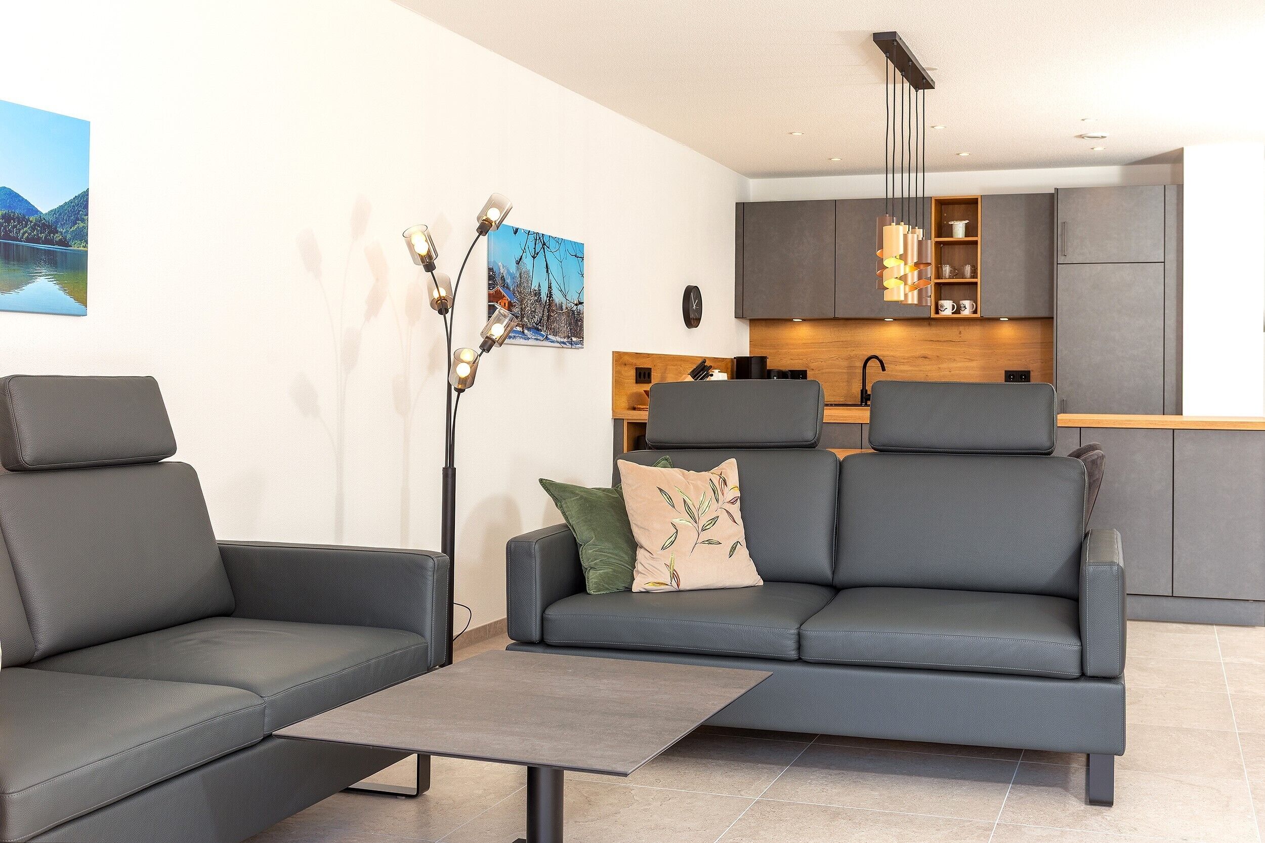 Una sala moderna con un sofá gris, una mesita de centro y una lámpara de pie. Se ve una cocina con gabinetes grises y una encimera de madera al fondo.