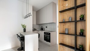 Uma cozinha moderna com armários brancos, uma ilha central com banquetas, forno embutido e uma estante de madeira com plantas.