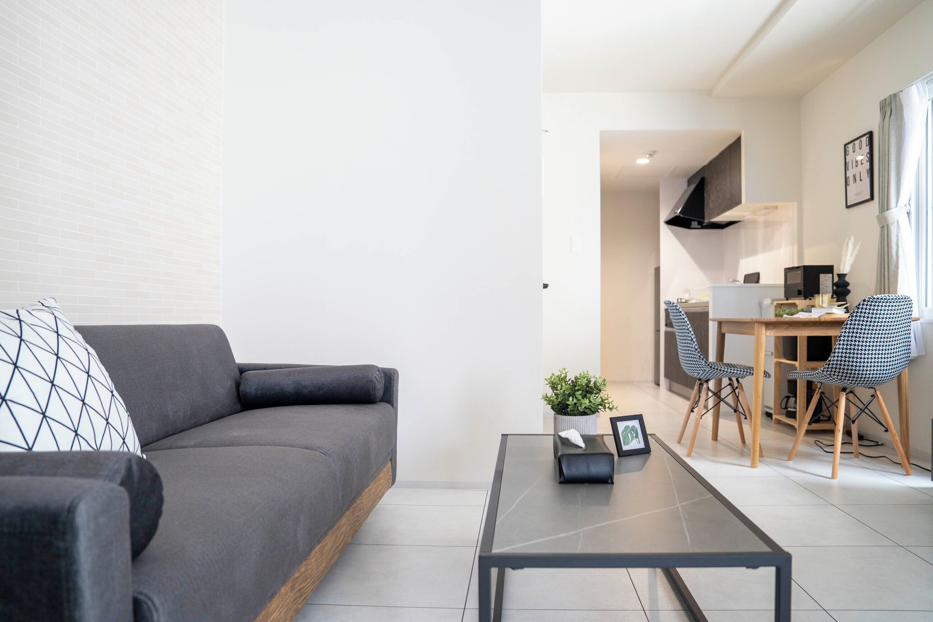 A modern living room with a grey sofa, a small table with a plant, and a dining area with a wooden table and chairs.