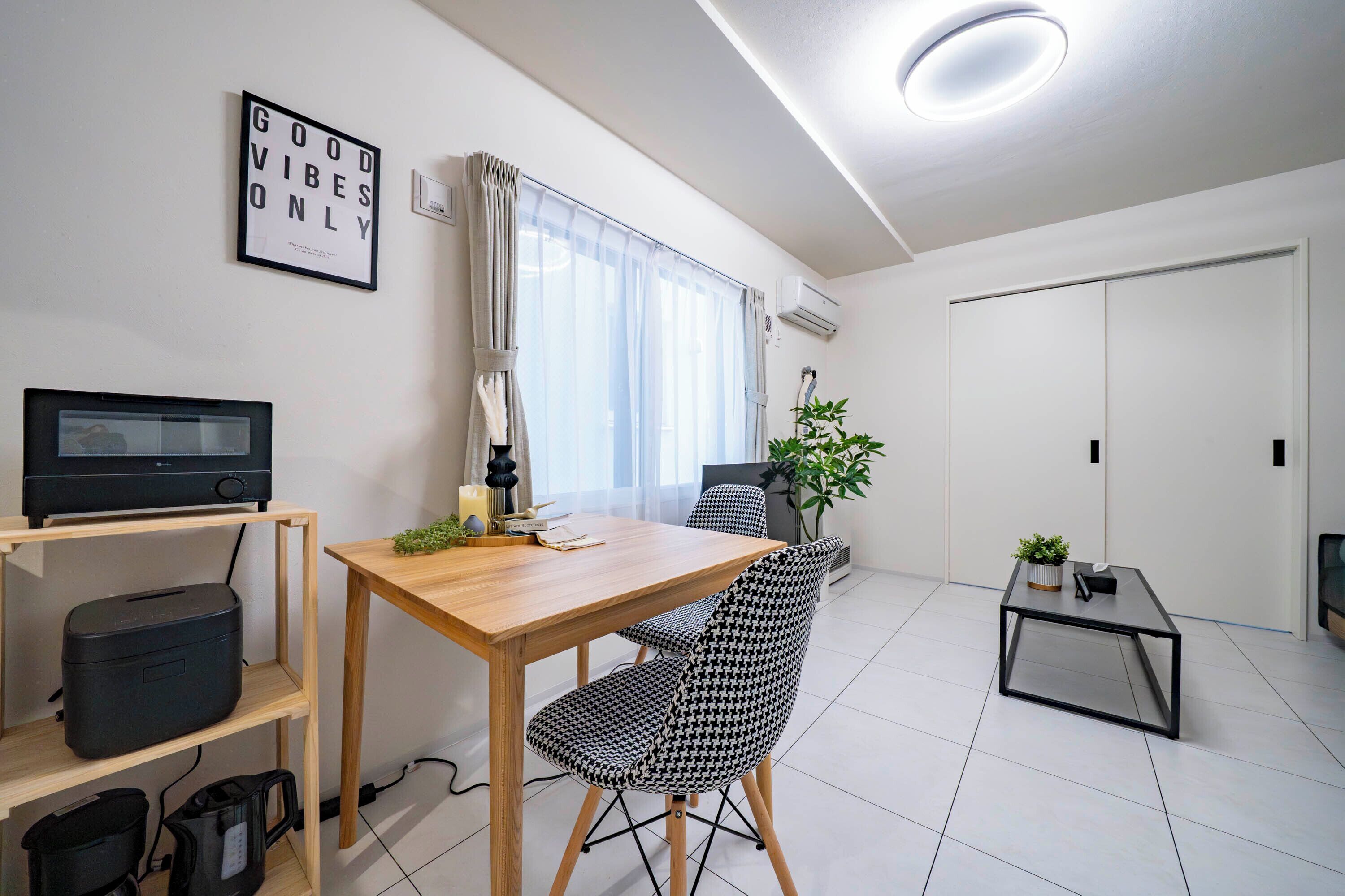 Une pièce moderne et bien éclairée, avec une table à manger en bois, deux chaises à motifs, une petite table d’appoint, un téléviseur posé sur une étagère et une plante en pot.