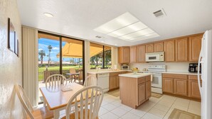 A kitchen with wooden cabinets, a microwave, an oven, and a dishwasher. There is a dining area with a table and chairs, and a view of a golf course through large windows.