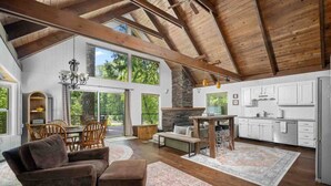 A spacious living area with a high ceiling, wooden beams, a fireplace, a dining area, and a kitchen with white cabinets.