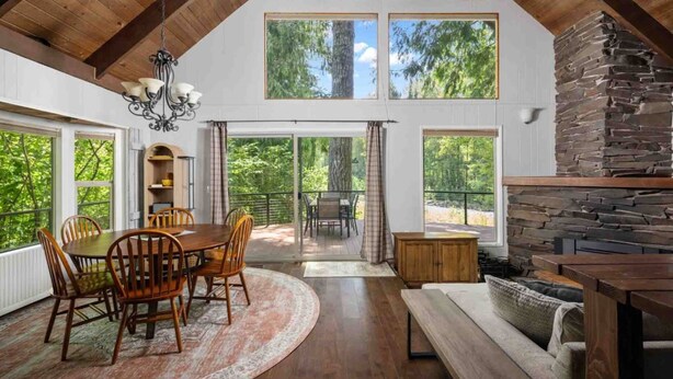 A spacious dining area with a wooden table and chairs, a stone fireplace, and large windows offering a view of the surrounding forest.