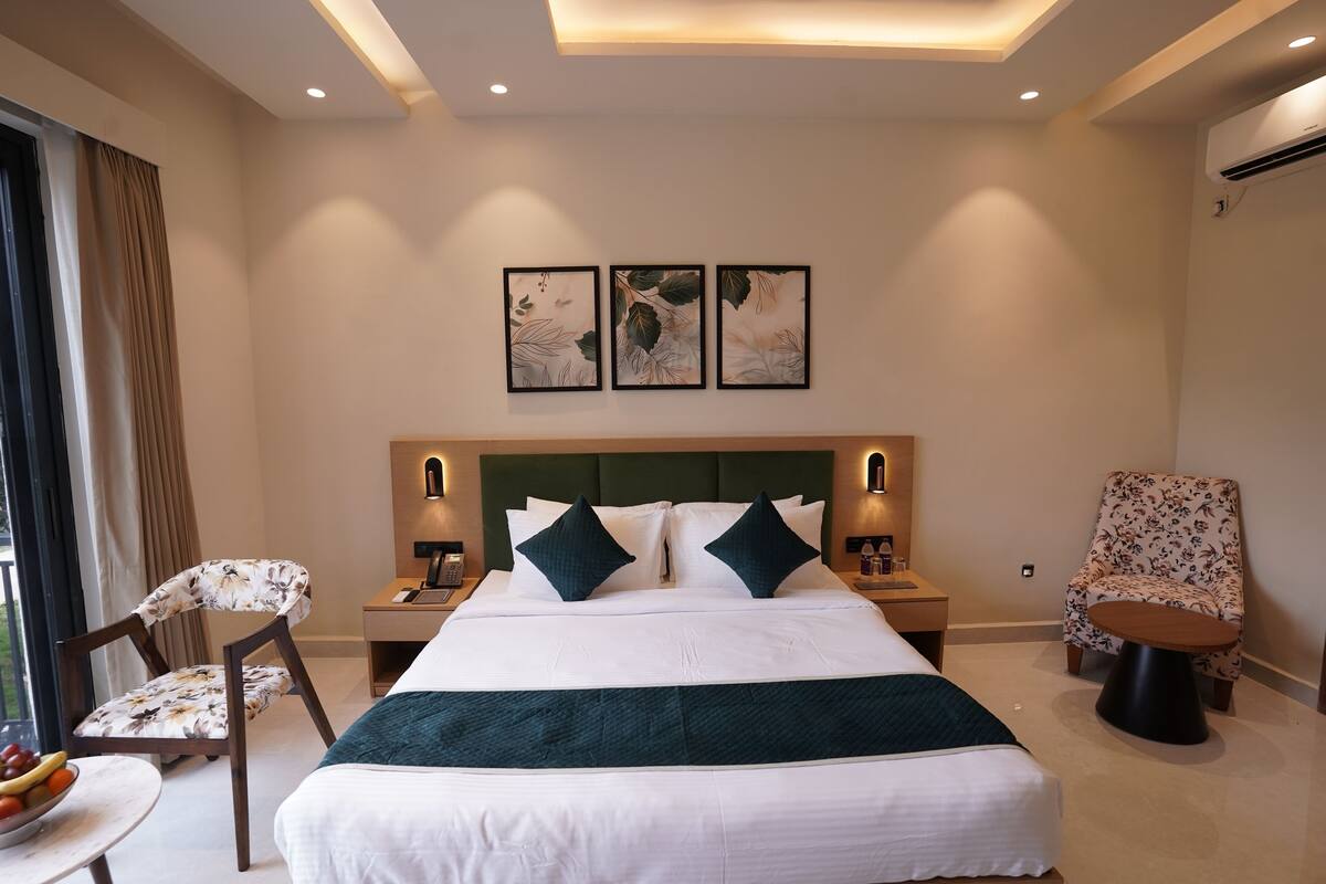 Una habitación de hotel moderna con una cama grande, dos mesitas de noche con lámparas, una silla, una pequeña mesa redonda con fruta y una ventana con cortinas.