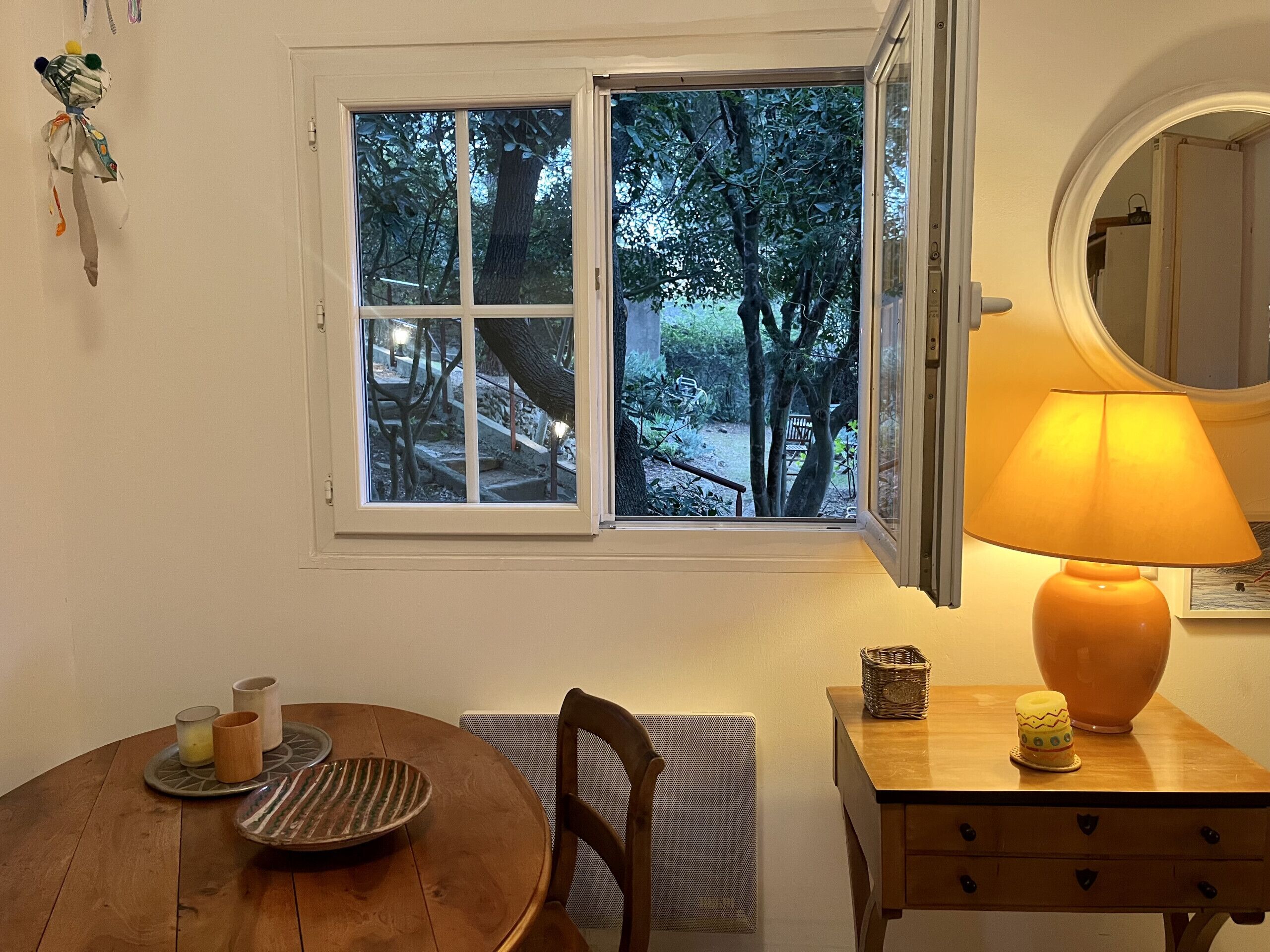 A room with a wooden dining table, a chair, a lamp, a small cabinet, and a window with a view of trees and a pathway.