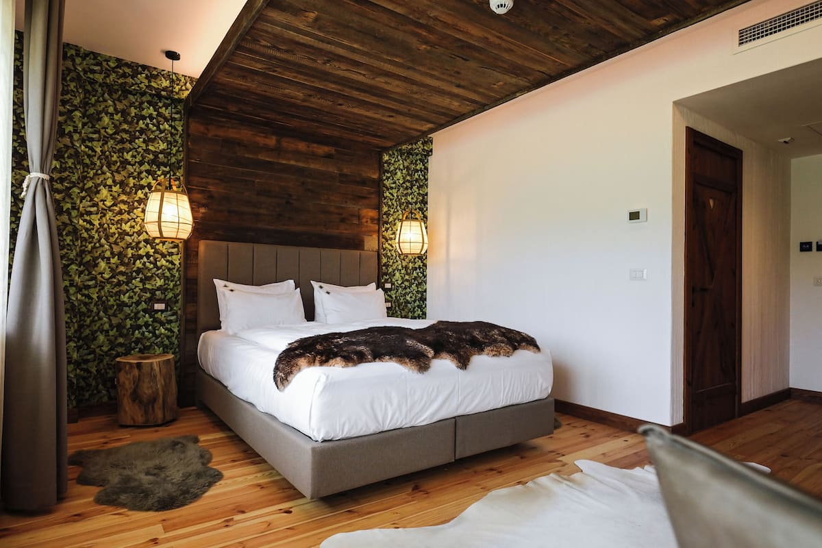 Une chambre à coucher avec un lit, un plancher en bois, un plafond avec des poutres apparentes et des murs recouverts de feuillage vert.