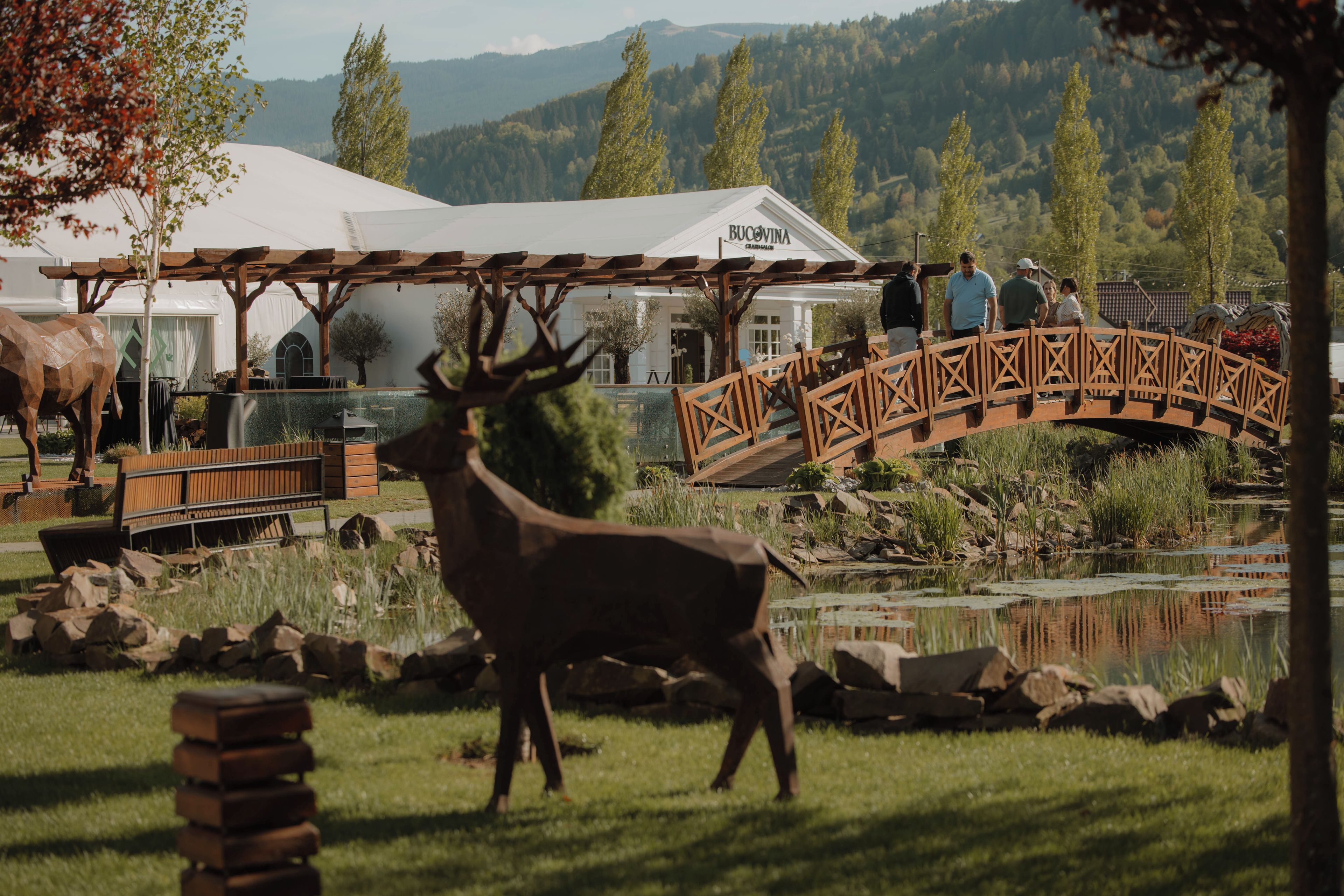 A rustic outdoor setting with a wooden bridge, a deer sculpture, and a pond.
