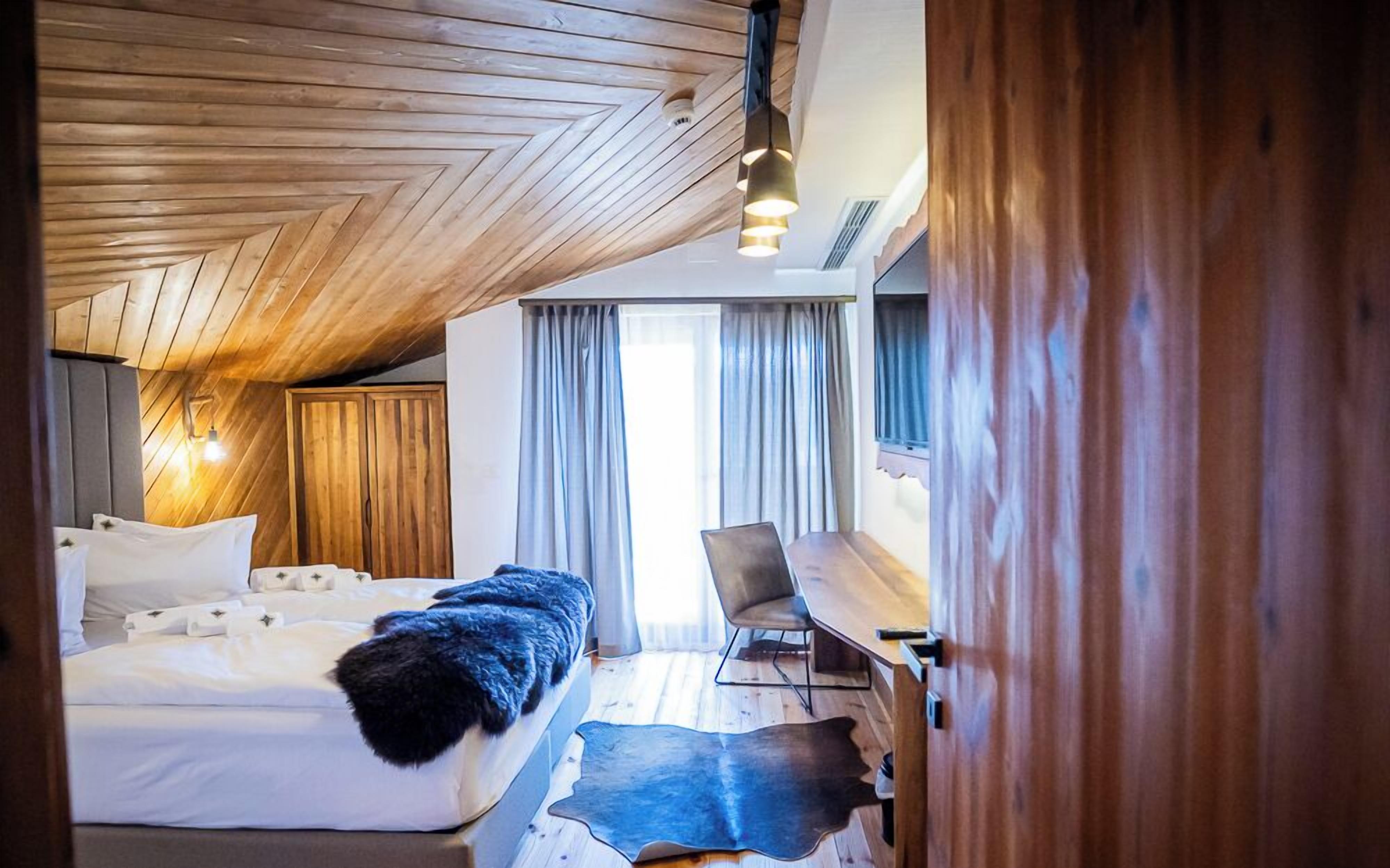 A bedroom with a wooden ceiling, a bed with white linens, a fur throw, a leather rug, a desk, and a chair.