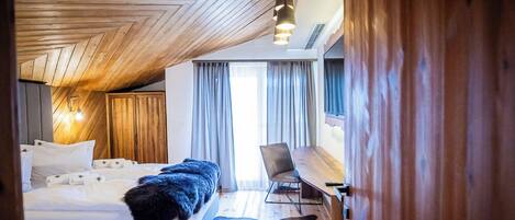 A bedroom with a wooden ceiling, a bed with white linens, a fur throw, a leather rug, a desk, and a chair.