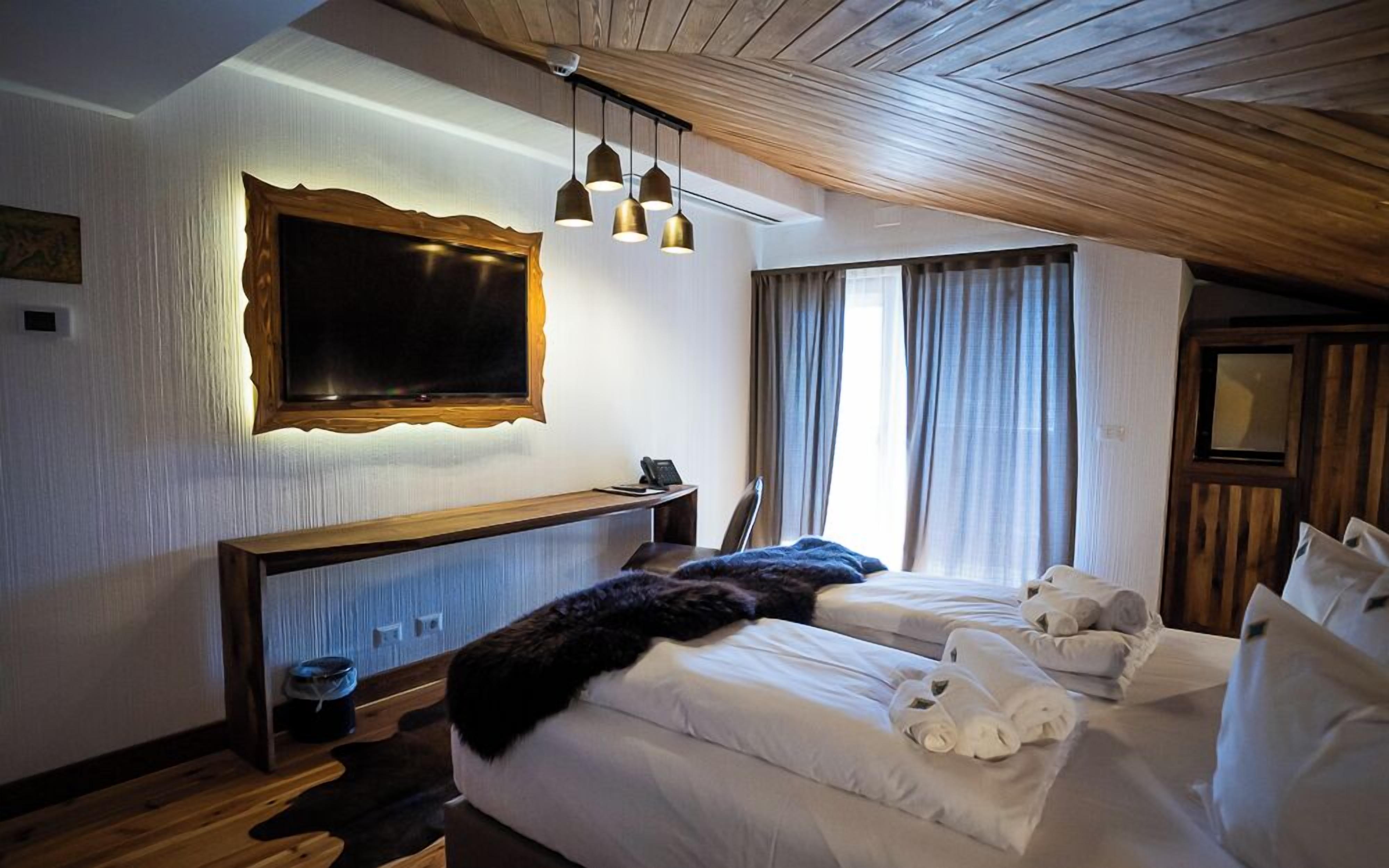 A hotel room with a large bed, bedside tables, a desk, a chair, a TV, and a window with curtains.