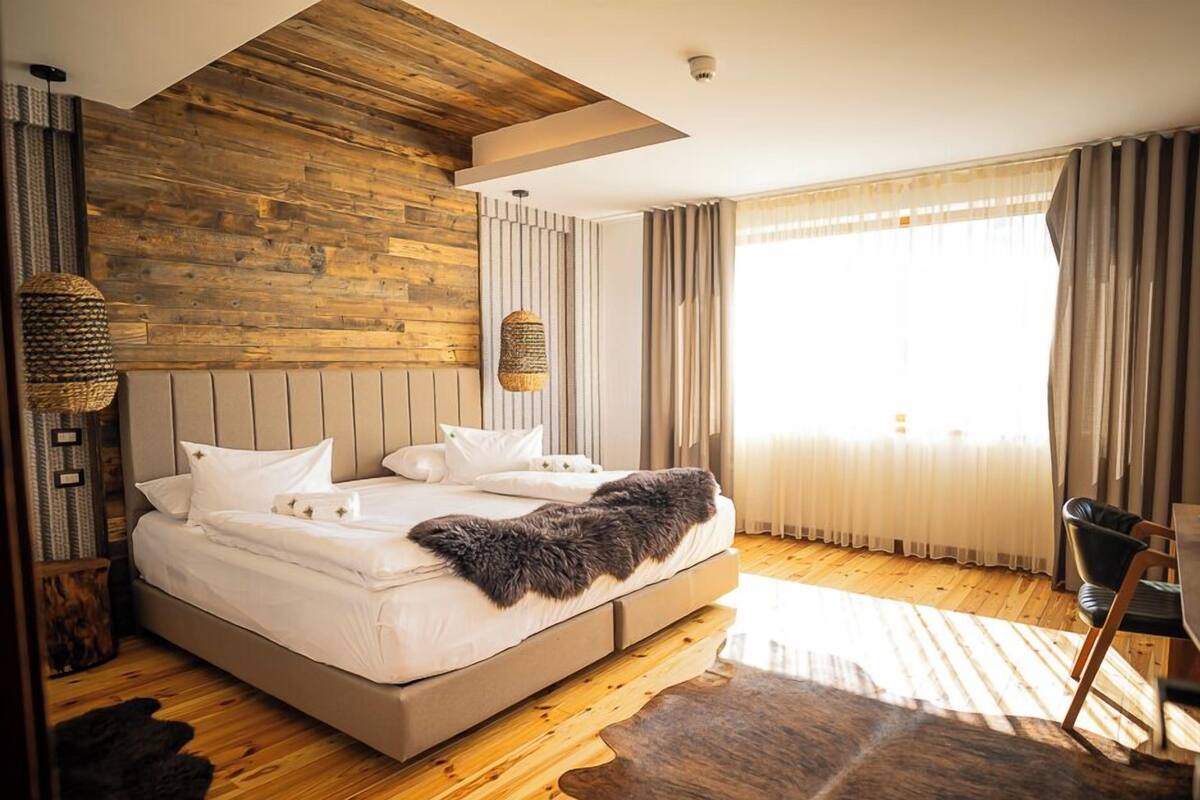 Une chambre moderne avec un plafond en bois, un grand lit recouvert de draps blancs, un plaid en fausse fourrure et une vue sur l’extérieur à travers des rideaux transparents.