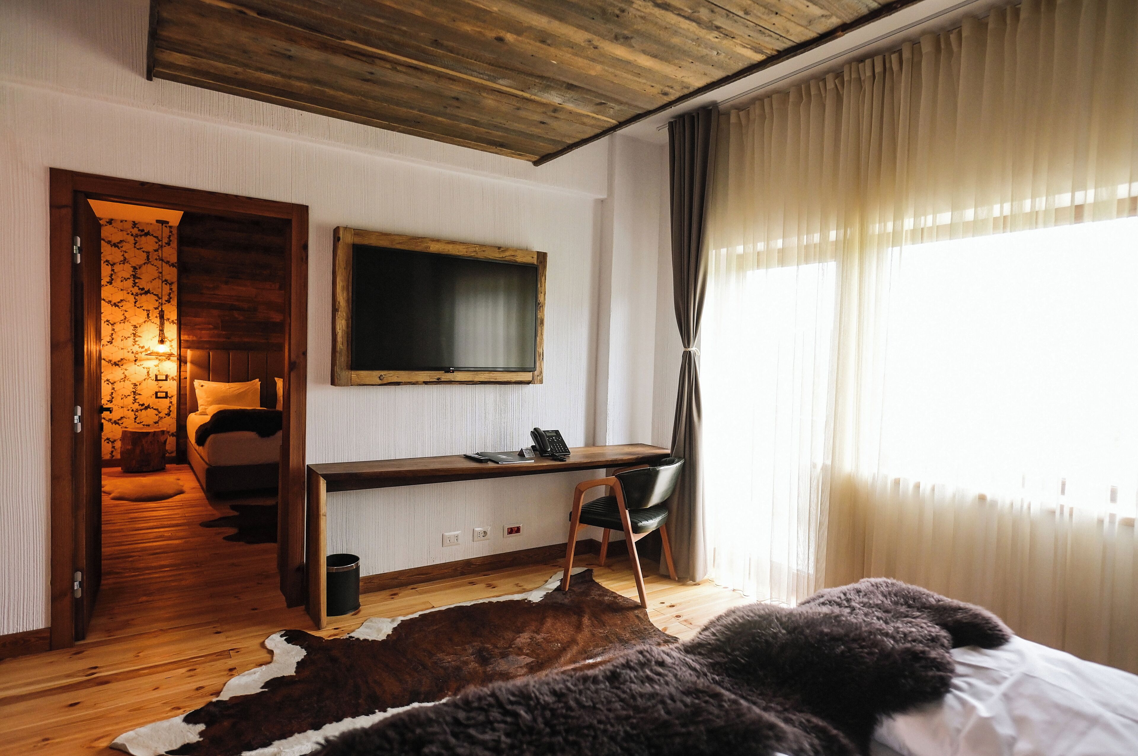 A room with a wooden desk, a chair, a bed with a large brown and white rug, a TV on the wall, and a window with sheer curtains.