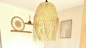 Un dormitorio con cama blanca, ropa de cama gris, mesita de noche de madera, lámpara colgante tejida y estante en la pared con objetos decorativos.