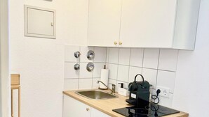 Cocina compacta con armarios blancos, un lavamanos de acero inoxidable, una cafetera y un cubo de basura.