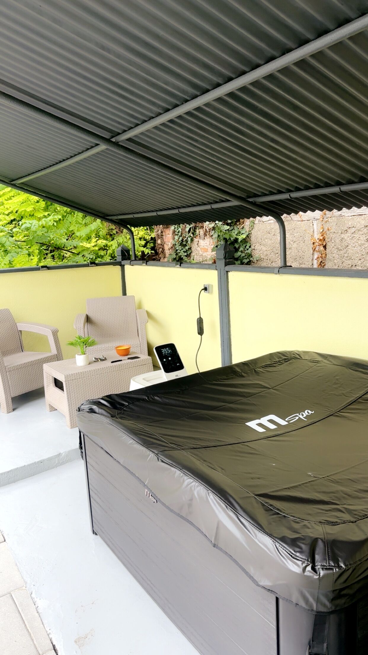 Een overdekt terras met een spa-bubbelbad, rieten meubilair en een bakstenen muur.