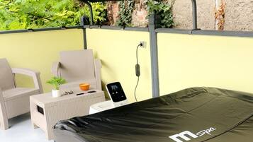Een overdekt terras met een spa-bubbelbad, rieten meubilair en een bakstenen muur.