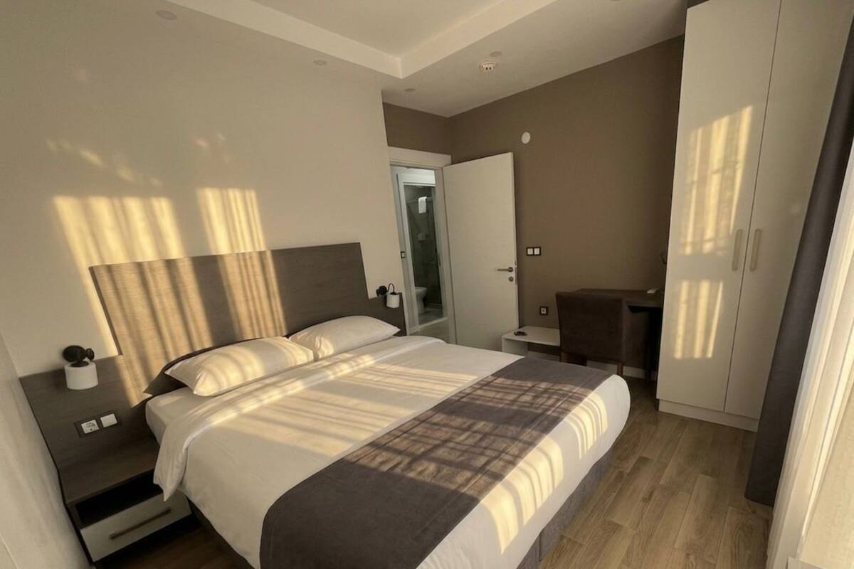 A modern hotel room with a large bed, bedside tables, a wardrobe, and a bathroom visible through an open door.