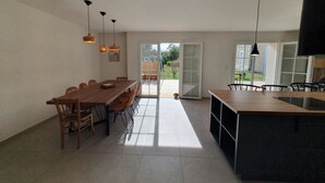 A spacious dining area with a long wooden table and chairs, a kitchen island, pendant lights, and a sliding glass door leading to an outdoor patio. - Sunny vacation home. Ideally located between lake, beach and forest. (Sanguinet)
