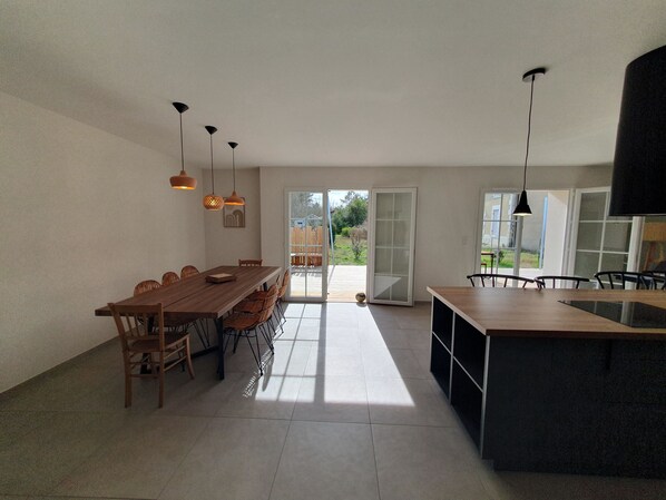 A spacious dining area with a long wooden table and chairs, a kitchen island, pendant lights, and a sliding glass door leading to an outdoor patio.