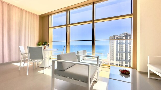 Quarto moderno com uma grande janela, mobiliário branco e vista para a cidade e oceano.