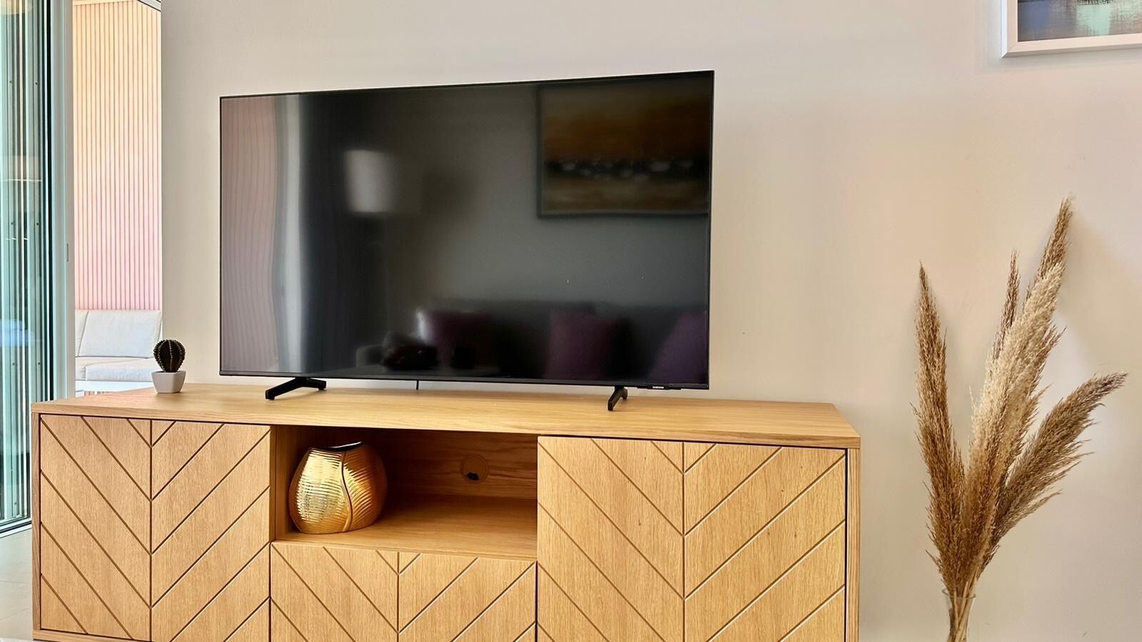 A flat-screen TV on a wooden TV stand with geometric patterns, a vase, and pampas grass.