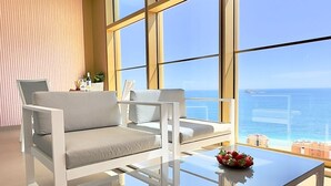 Uma sala de estar moderna com uma mesa de centro de vidro, dois sofás brancos e vista para o mar atrav és de amplas janelas.