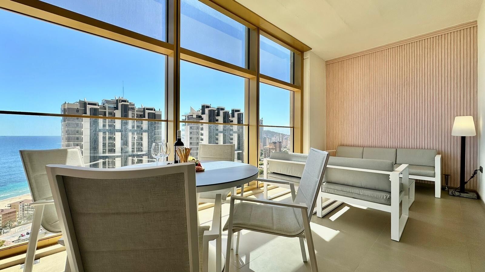 Habitación de hotel moderna con un ventanal grande que ofrece vistas a la ciudad, una mesa redonda con sillas, un sofá y una lámpara.