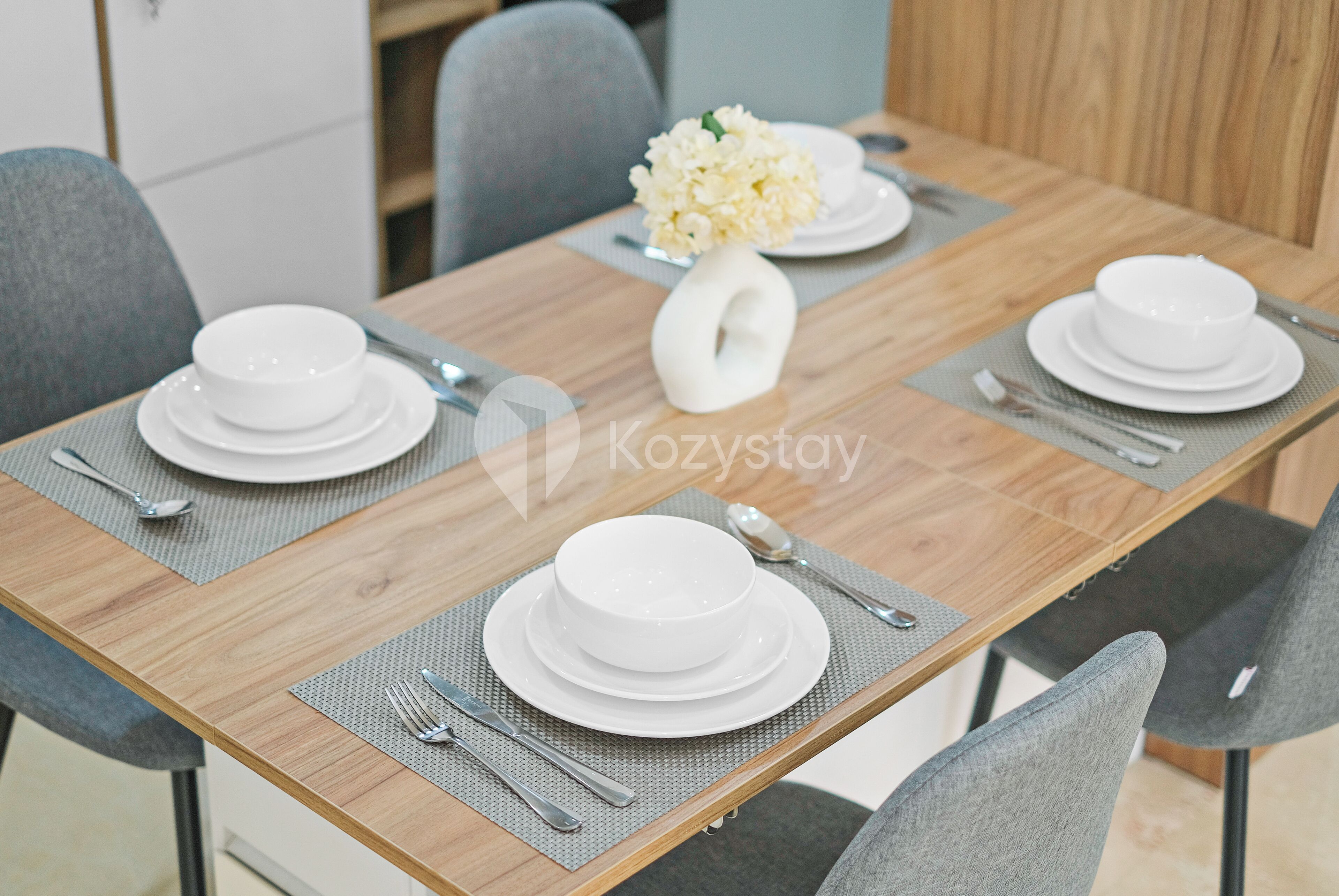 A dining table set with white plates, bowls, and cutlery, with a white vase containing white flowers in the center.