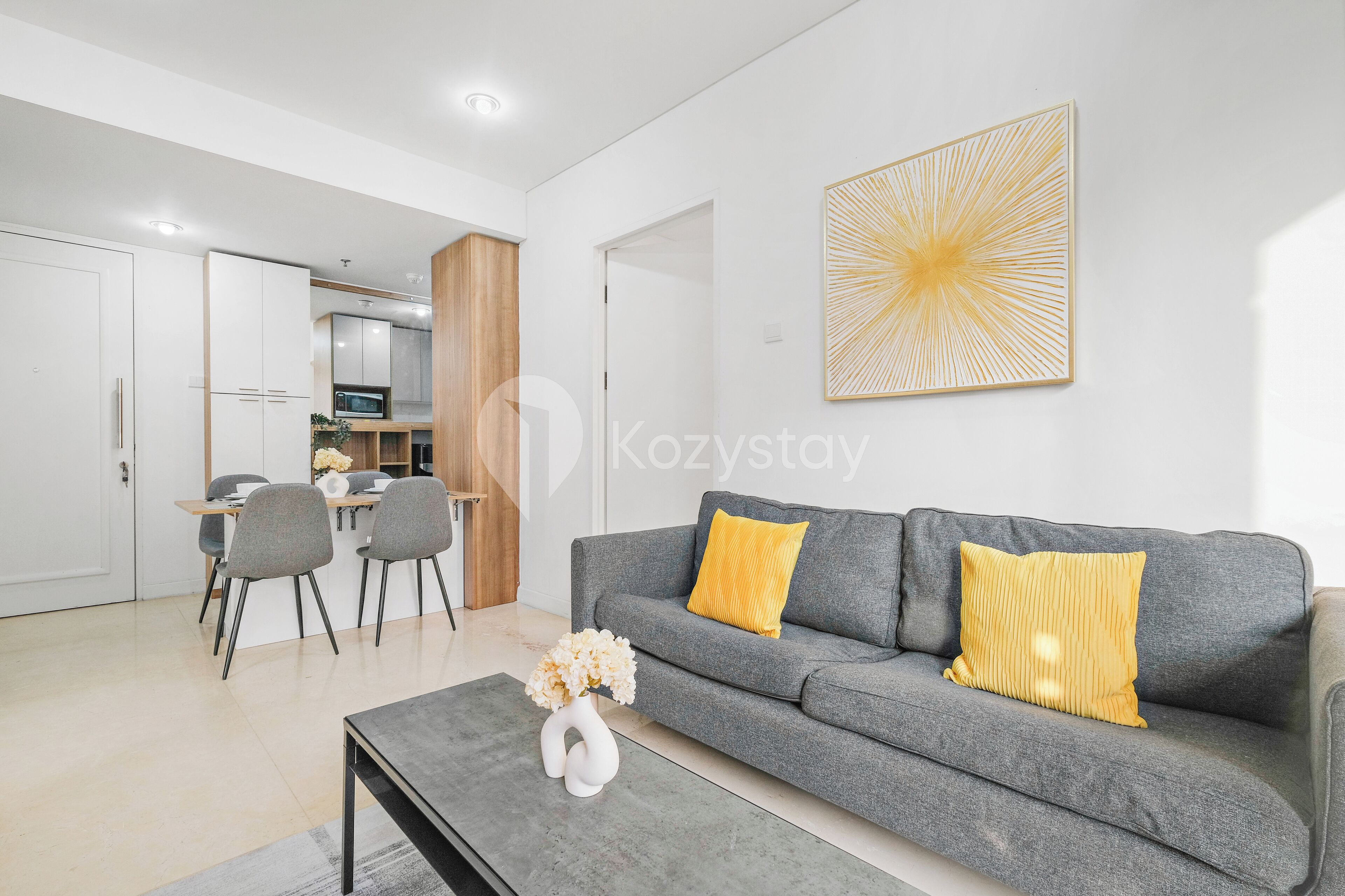 Una sala moderna con un sofá gris, dos cojines amarillos, un jarrón blanco con flores y un comedor con mesa y sillas.
