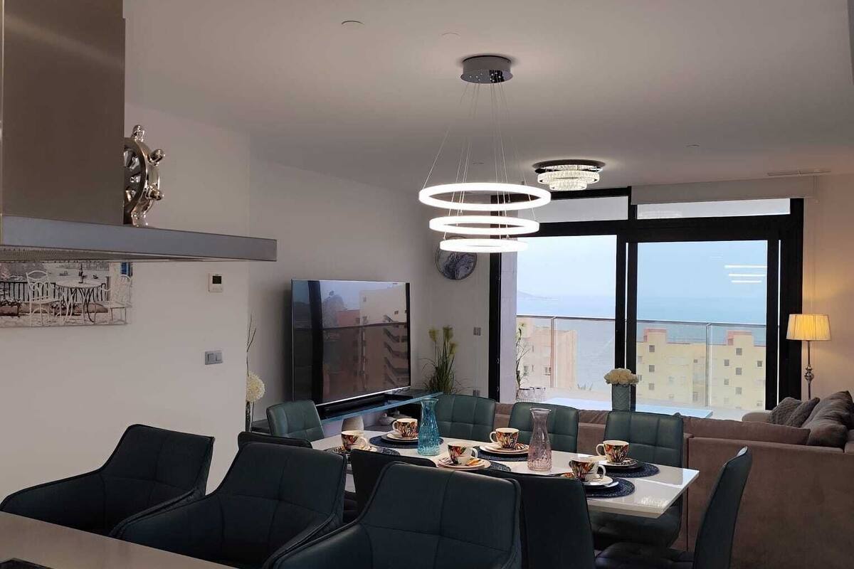 A modern kitchen with a dining area, a large window with a city view, and a living area with a sofa and lamp.