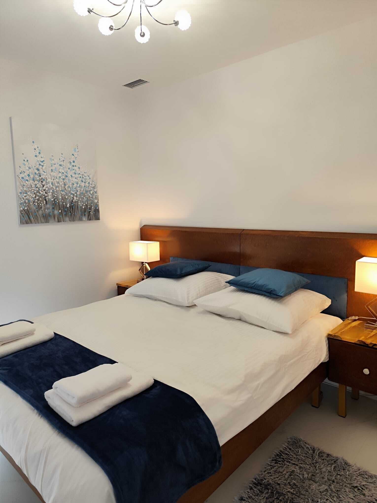 A neatly made bed with white and blue bedding, a wooden headboard, and a wall-mounted artwork.