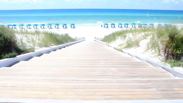 Un muelle de madera que conduce a una playa con sombrillas azules y sillas de playa blancas.