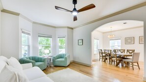 Un salón espacioso con un sofá blanco, sillones azul turquesa, una zona de comedor con una mesa y sillas de madera, y ventanas grandes con persianas.