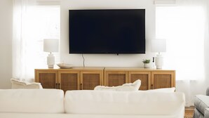 Una sala moderna con un televisor de pantalla plana grande montado en la pared, un mueble de madera con cestas de mimbre y mobiliario blanco.