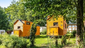 Dos cabañas de madera con un pequeño ventanal y puerta, rodeadas de vegetación y árboles.