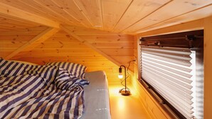 Habitación tipo cabaña de madera con cama, ventana con persianas e iluminación en la pared.