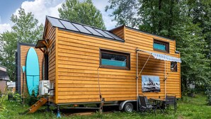 Un camping-car en bois doté d’une terrasse couverte, d’une planche de surf et d’une petite table avec des chaises.