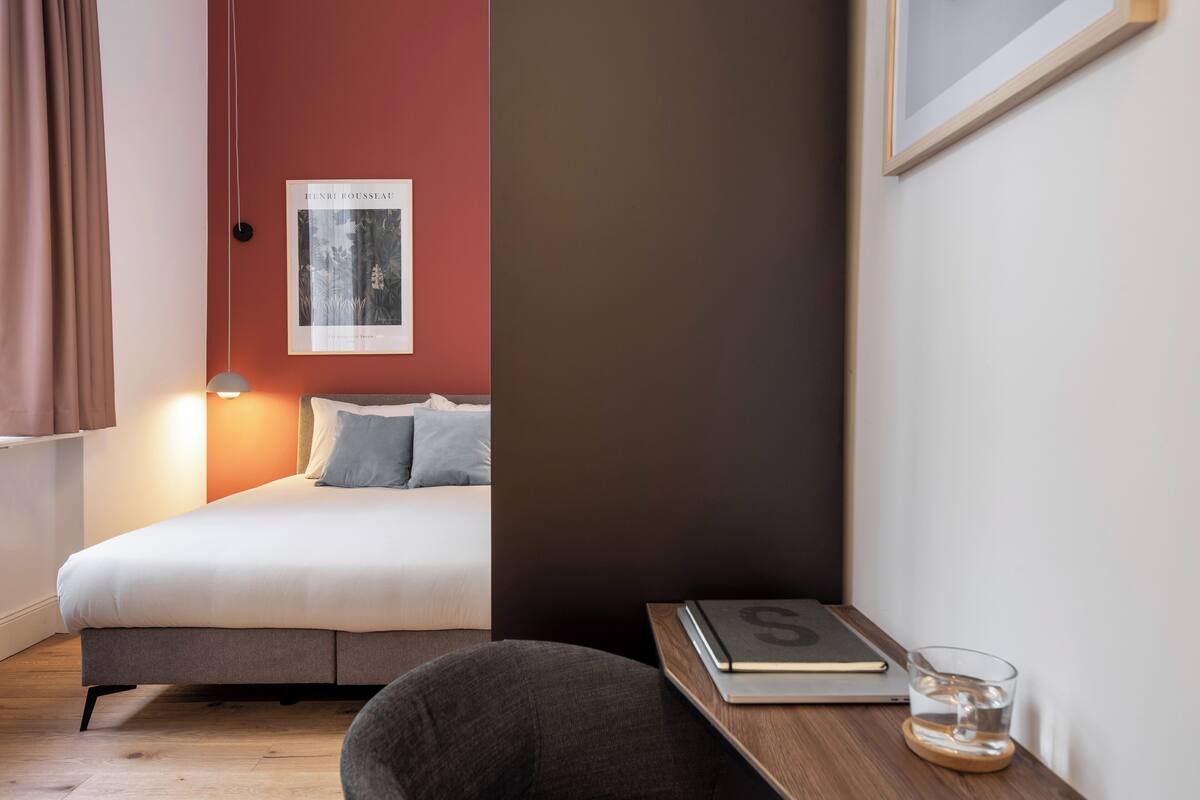 Ein modernes Schlafzimmer mit einem Bett, einem Nachttisch mit Buch und Glas, einem Poster an der Wand und einem Fenster mit Vorhängen.