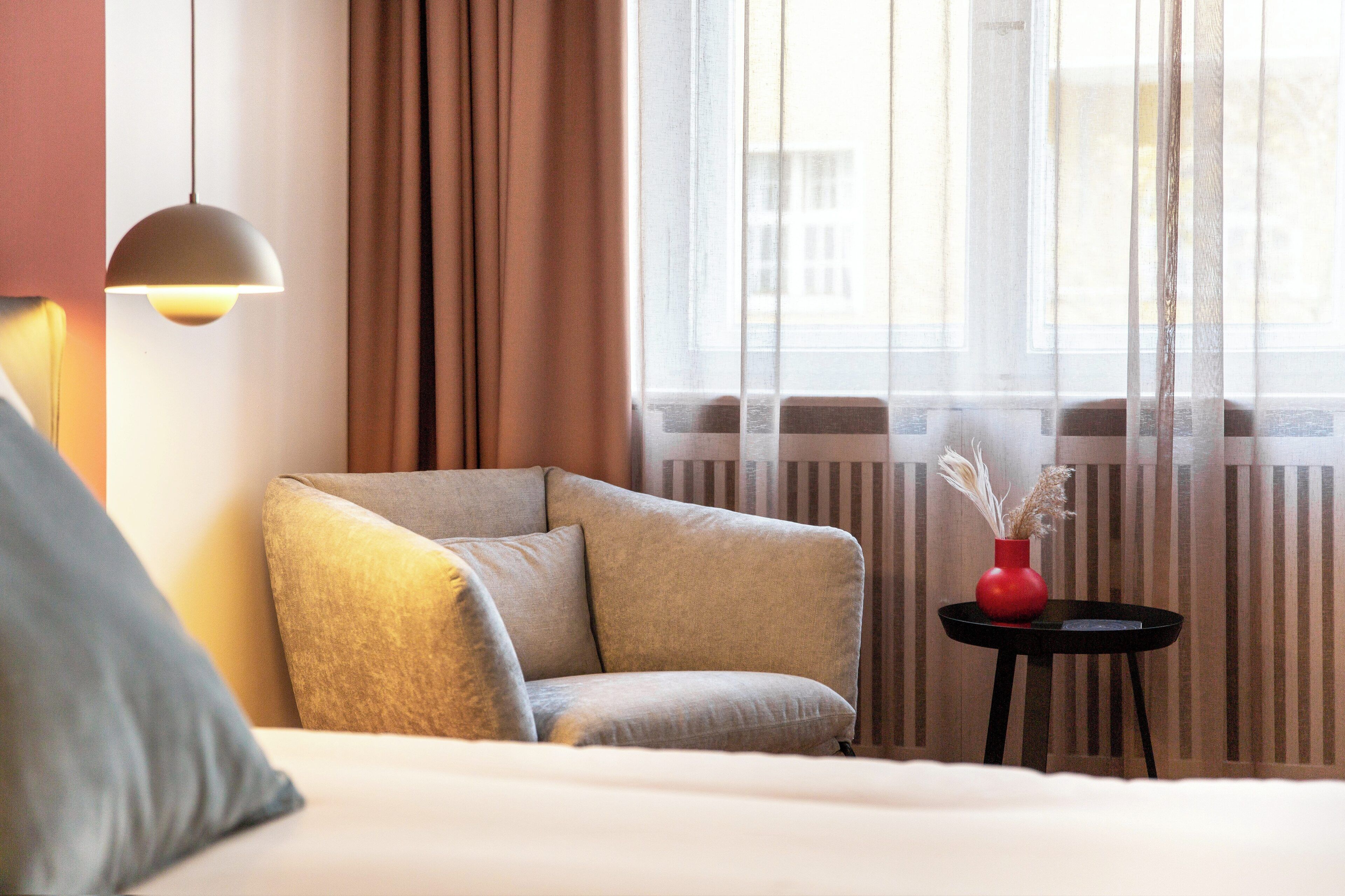 A modern hotel room with a sofa, a small round table with a red vase, and a hanging light fixture.