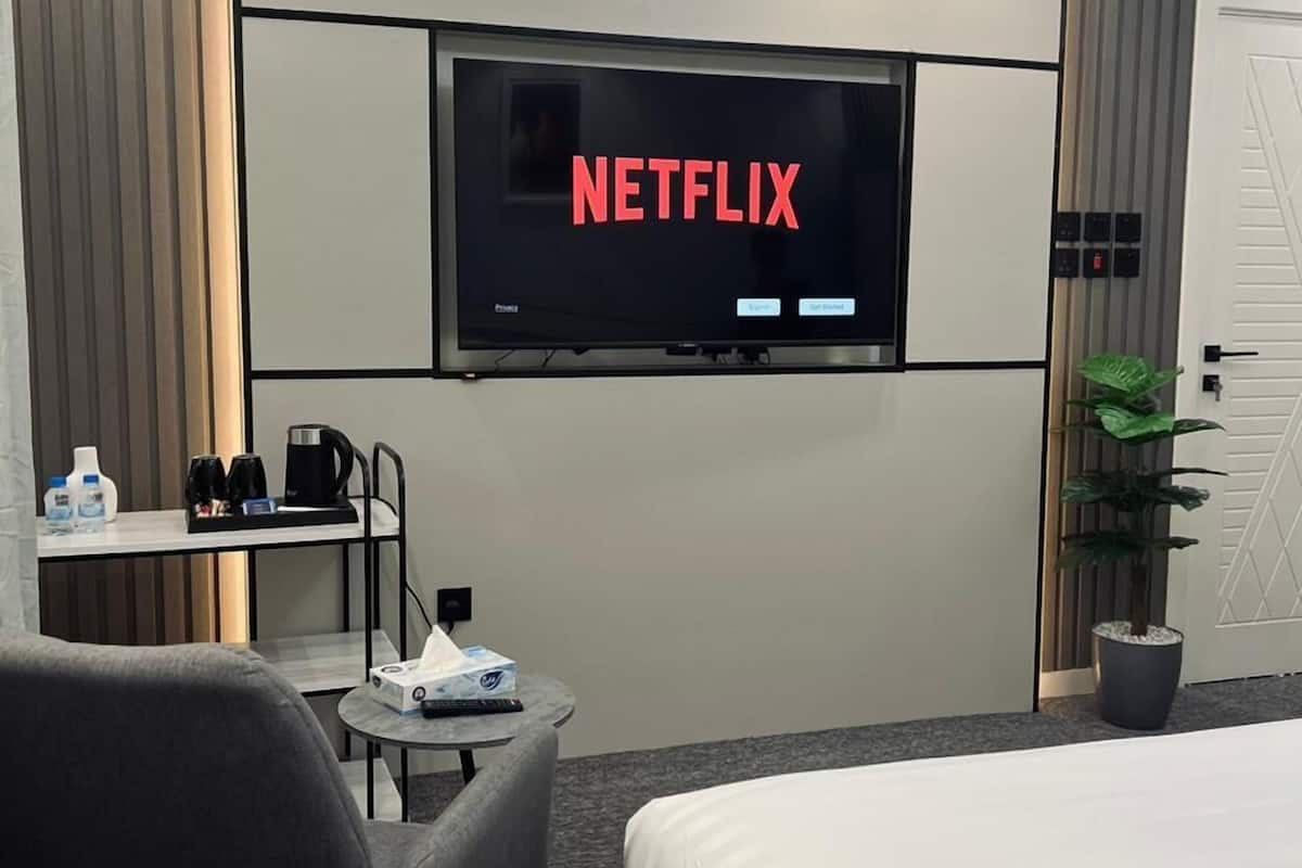 A hotel room with a large flat-screen TV mounted on the wall displaying the Netflix logo, a bed, a desk with a chair, a small table with a coffee maker, and a potted plant.