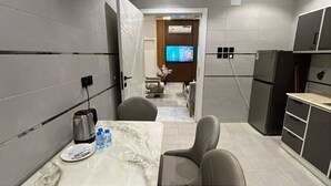 A compact hotel room with a marble table, two chairs, a small kitchenette, a TV, and a door leading to a hallway. - Downtown Suites Orbitnet (Madinah)