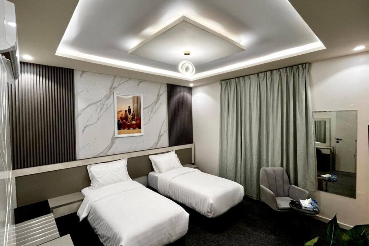 A modern hotel room with two single beds, a seating area with a chair, a small table, and a large mirror.