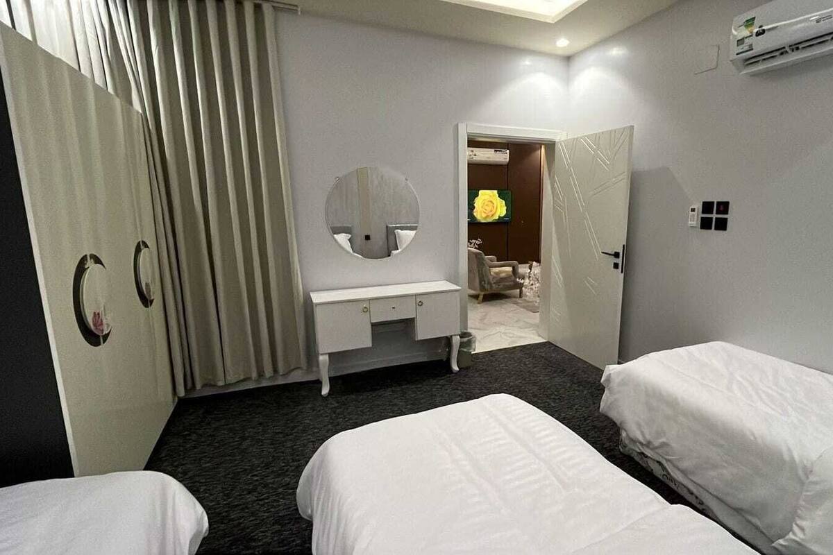 A hotel room with two single beds, a white dresser, a round mirror, a door with a light switch, and an air conditioning unit.