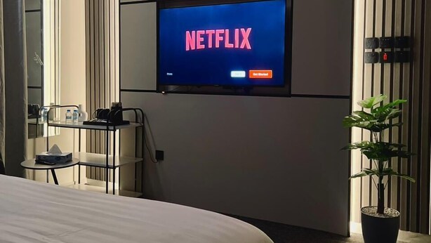A hotel room with a large bed, a TV mounted on the wall displaying 'NETFLIX', a small table with a chair, a potted plant, and a mirror. - Downtown Suites Orbitnet (Madinah)