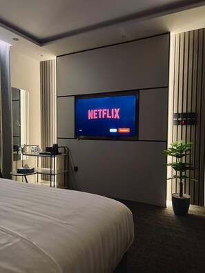 A hotel room with a large bed, a TV mounted on the wall displaying 'NETFLIX', a small table with a chair, a potted plant, and a mirror. - Downtown Suites Orbitnet (Madinah)