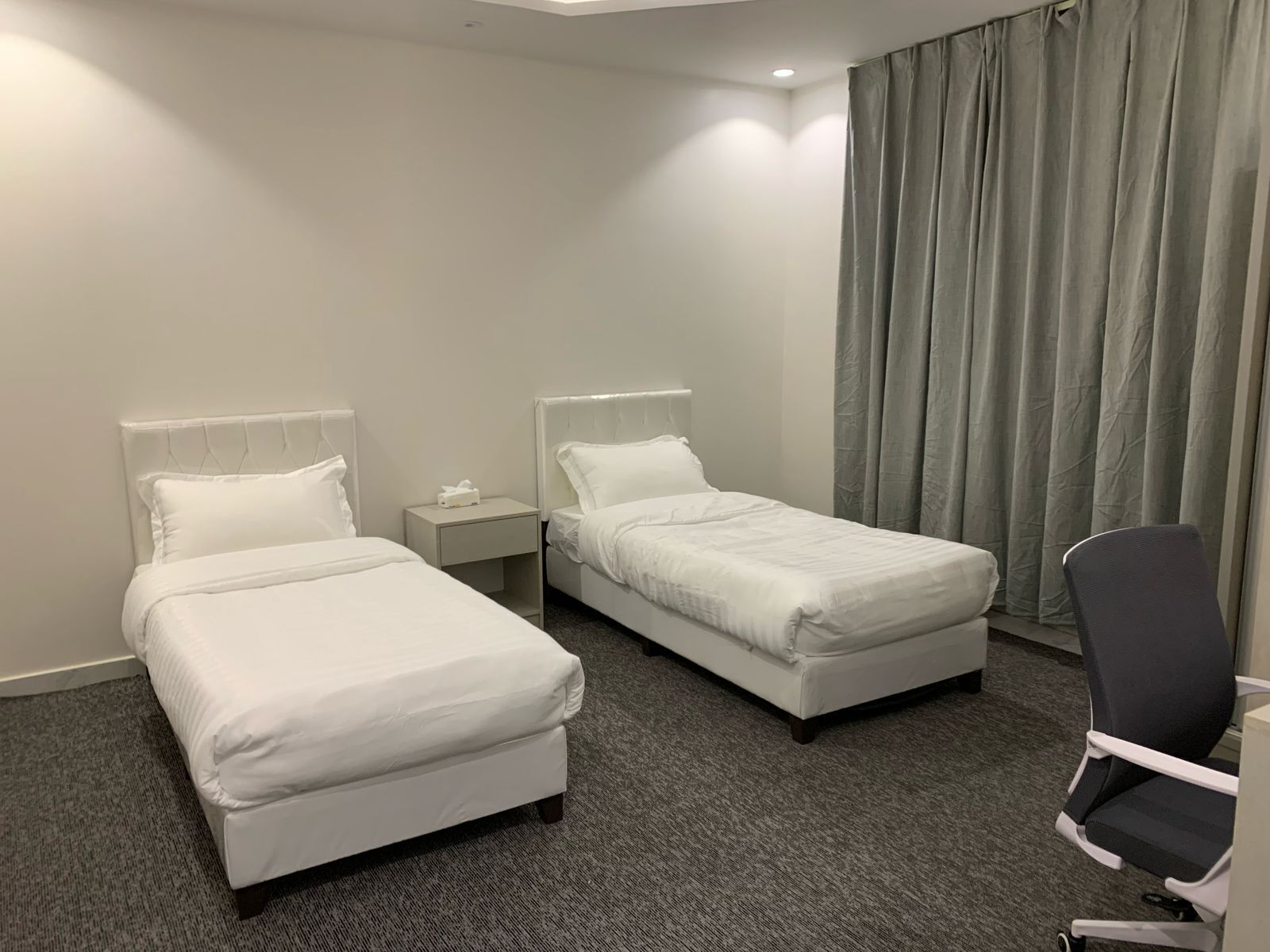 A hotel room with two single beds, a small bedside table with a lamp, a chair, and a large window with curtains.