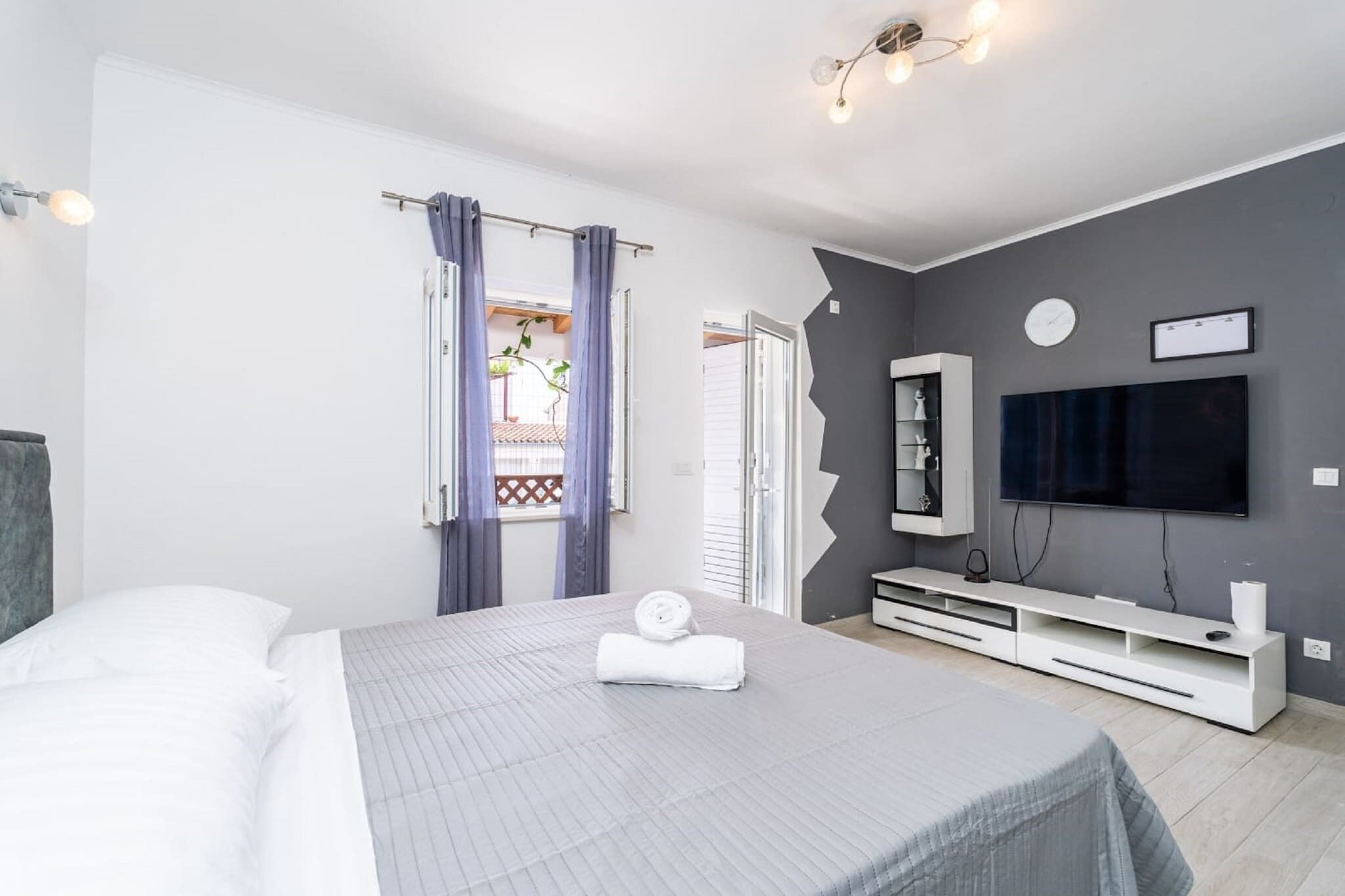 Quarto de hotel moderno com uma cama grande, televisão na parede e uma varanda com cortinas.