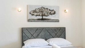 Een netjes opgemaakt bed met een grijs beklede hoofdbord, witte kussens en een opgerolde witte handdoek. Daarboven hangt een lijst met een afbeelding van een boom aan een effen muur.