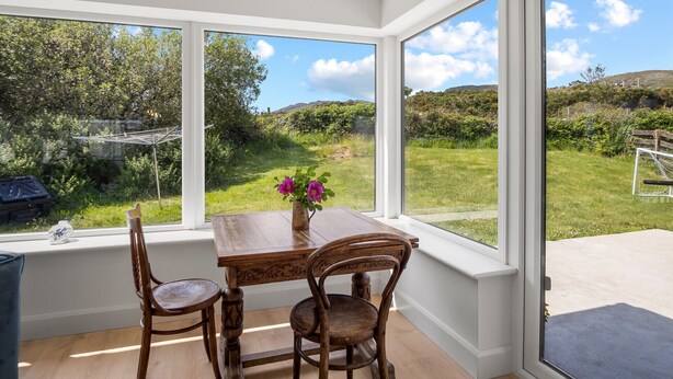 A dining area with a wooden table and chairs, large windows offering a view of a grassy landscape and hills.